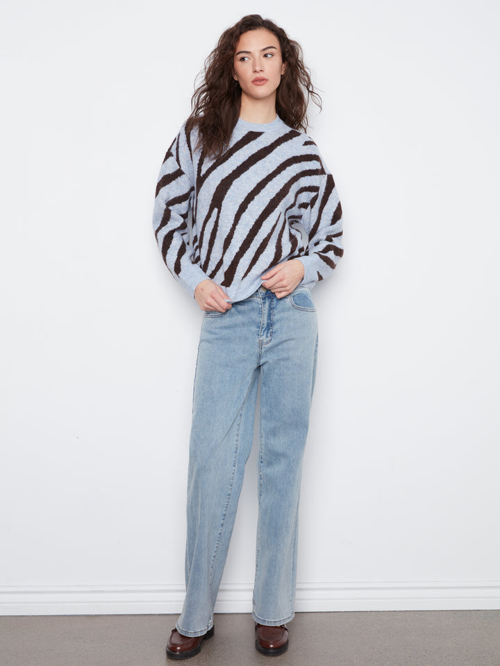A woman modeling a light gray and black zebra-striped knit sweater, paired with light-wash blue jeans and dark brown shoes, standing against a plain white wall.