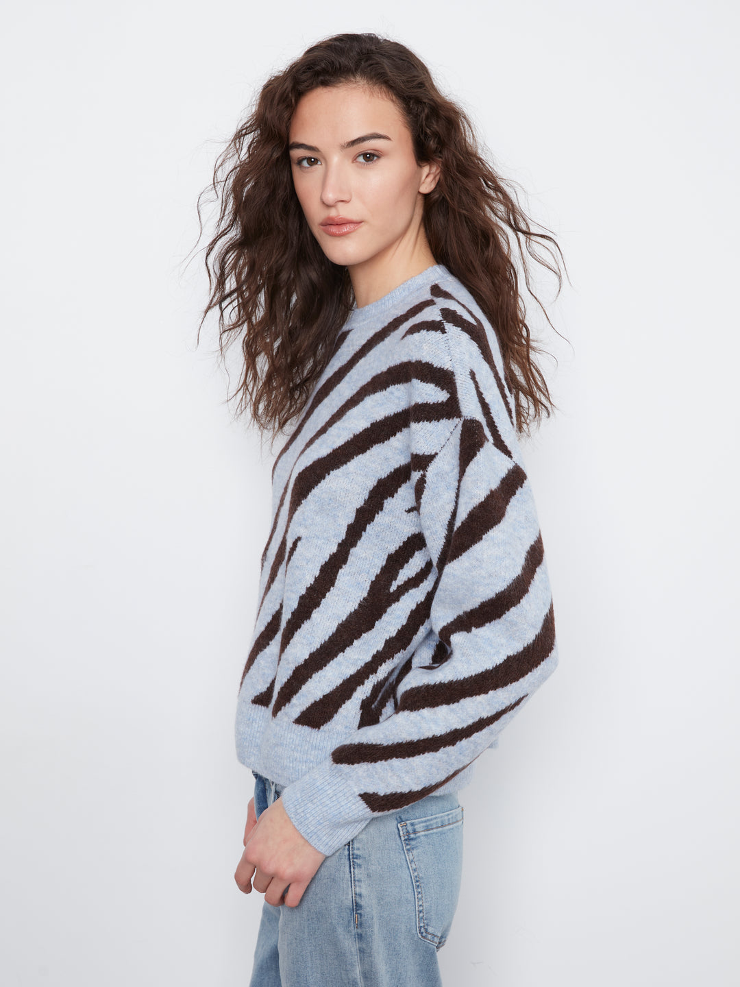 A woman with long, wavy brown hair is shown from the side, wearing a light gray fuzzy knit sweater with bold dark brown zebra-like stripes and light-wash blue jeans, set against a plain white studio background.