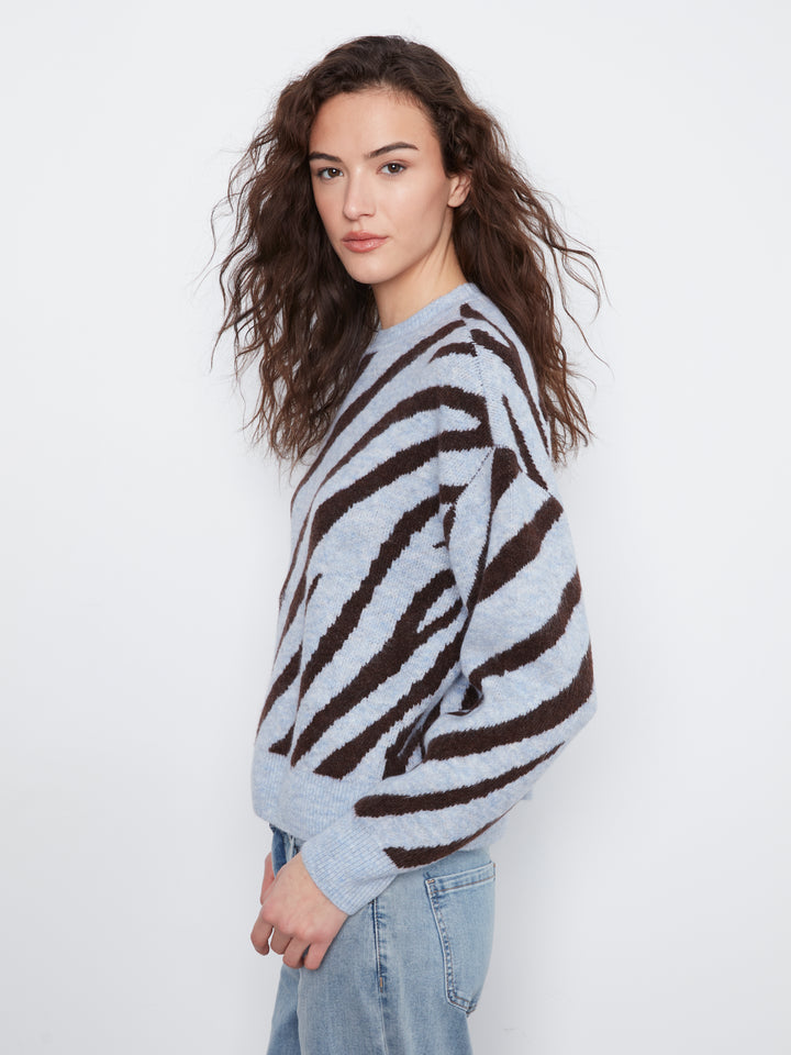 A woman with long, wavy brown hair is shown from the side, wearing a light gray fuzzy knit sweater with bold dark brown zebra-like stripes and light-wash blue jeans, set against a plain white studio background.