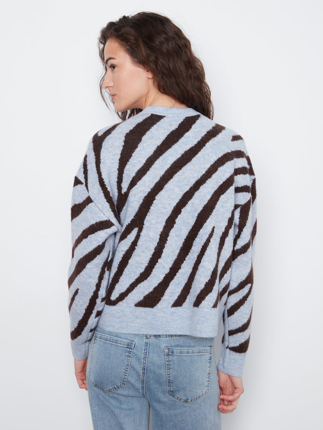A woman shown from the back wearing a light gray and dark brown zebra-striped knit sweater with light blue jeans, set against a plain white studio background.