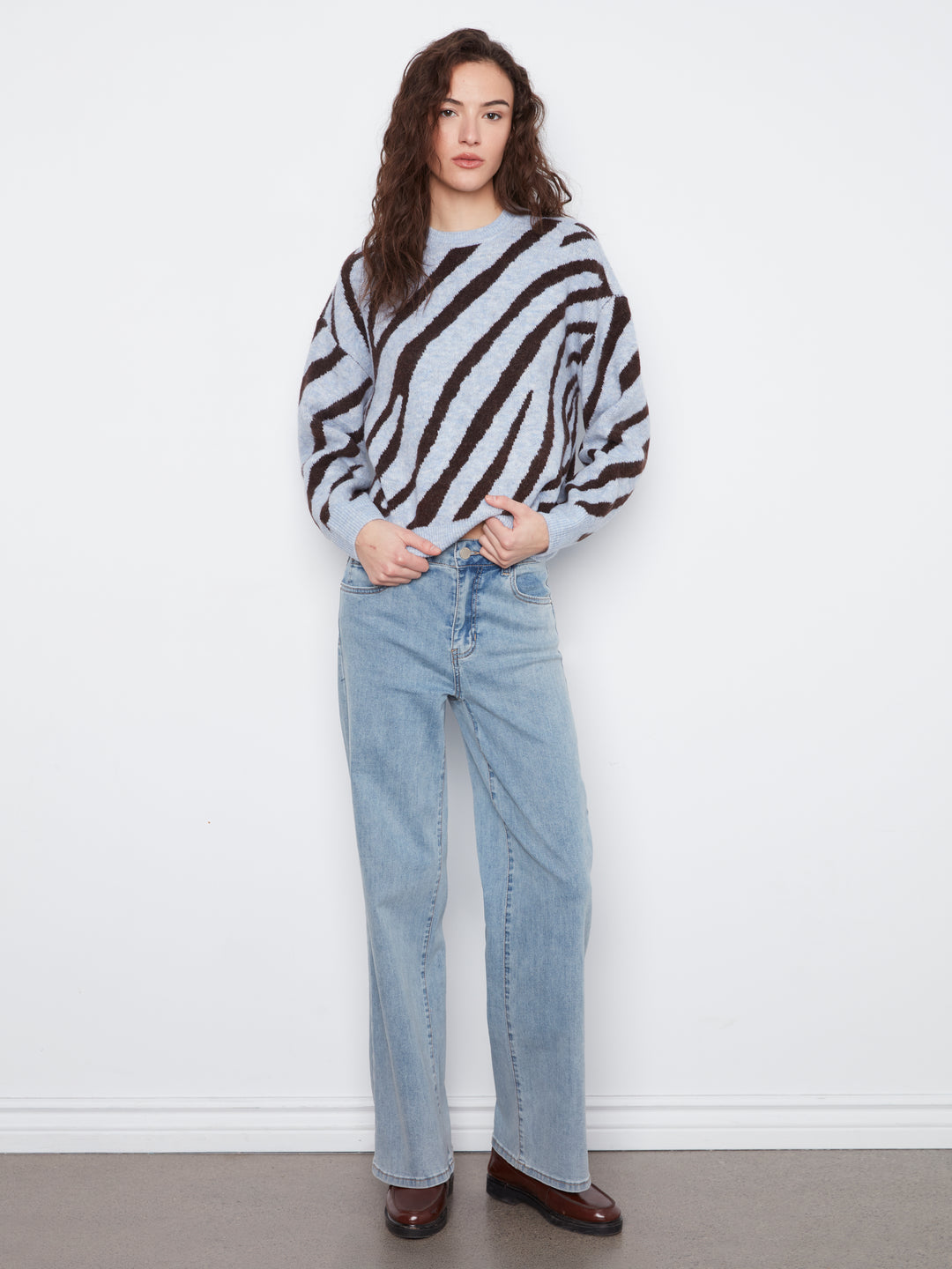 A woman stands against a white wall wearing a light gray knit sweater with bold black diagonal stripes, light-wash blue jeans, and dark brown shoes.