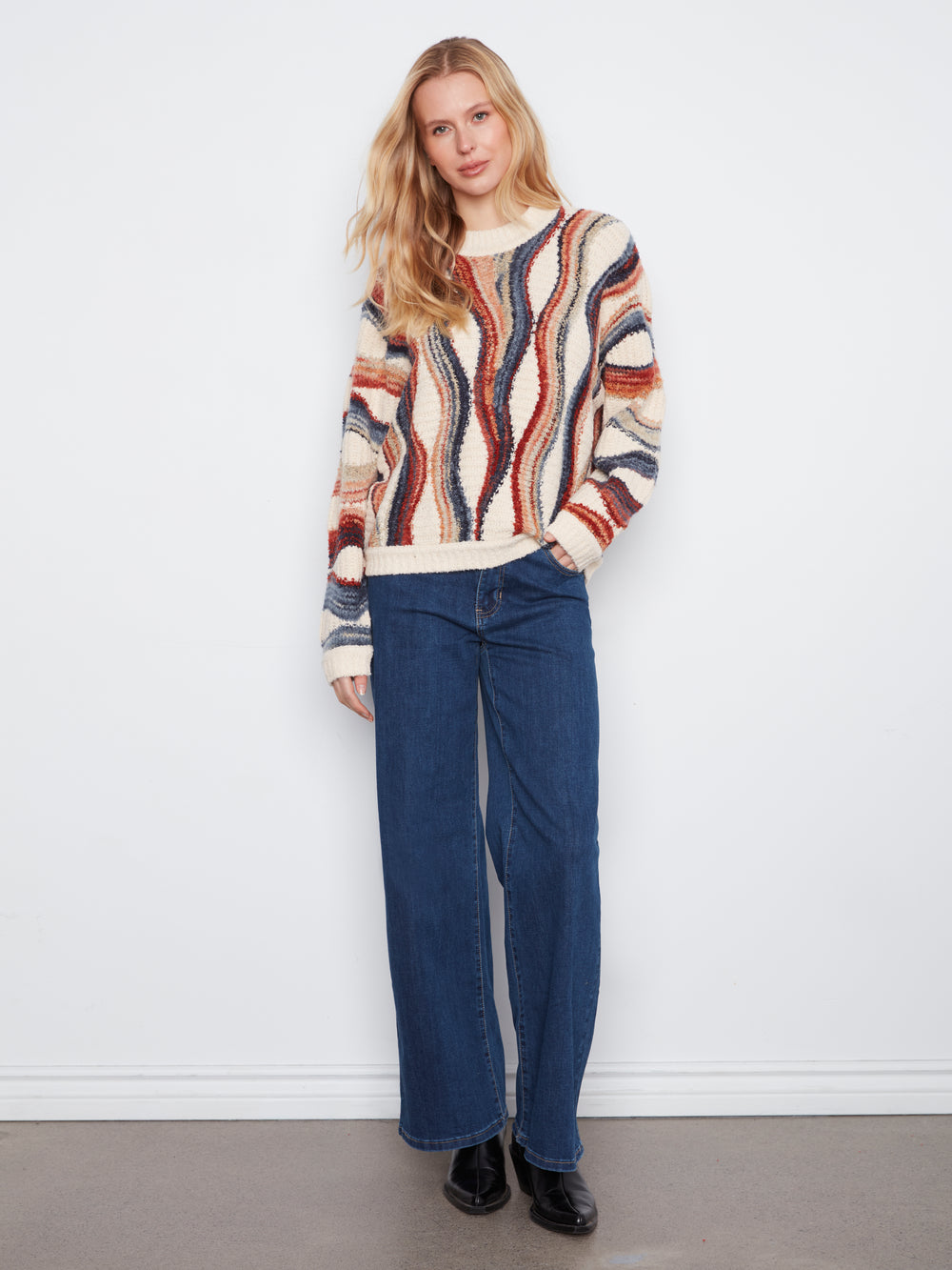 A woman stands against a white wall wearing a cream knit sweater with vertical wavy stripes in rust red, navy blue, and brown, paired with dark blue wide-leg jeans and black shoes.