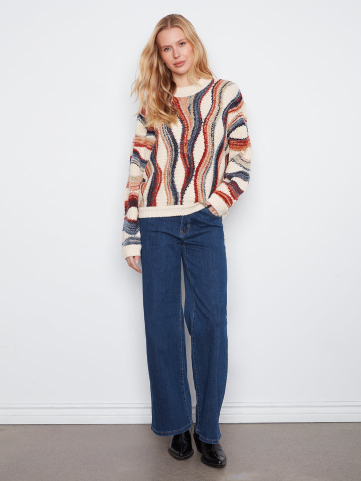 A woman stands against a white wall wearing a cream knit sweater with vertical wavy stripes in rust red, navy blue, and brown, paired with dark blue wide-leg jeans and black shoes.