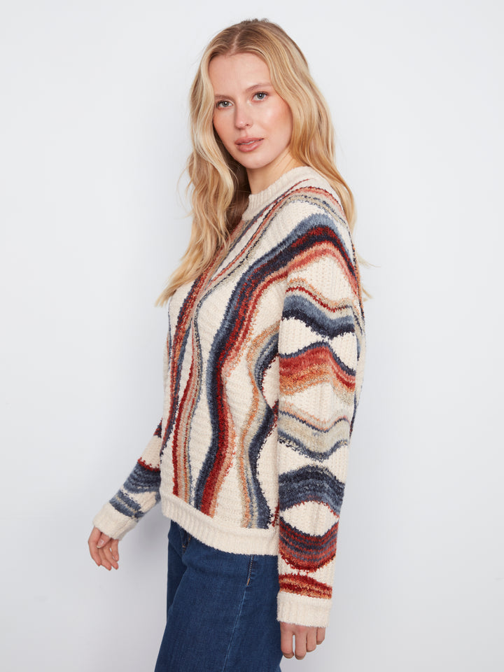 A woman modeling a chunky knit cardigan sweater with a wavy, multicolor striped pattern in cream, rust red, navy blue, and brown, paired with dark blue jeans against a plain light gray background.