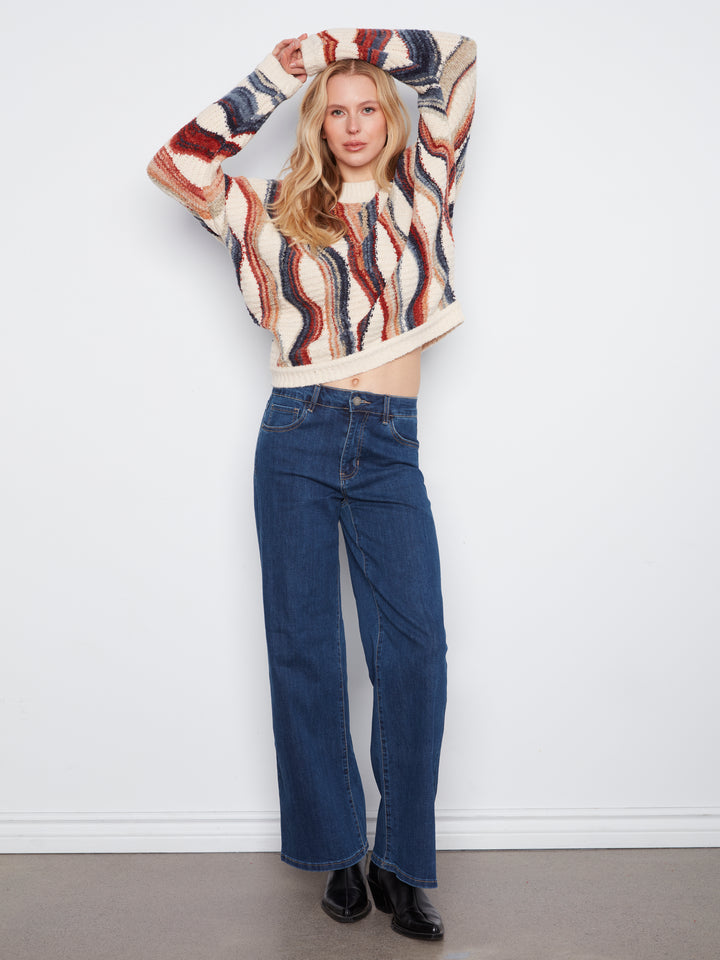 A woman modeling a cropped fuzzy knit sweater with a wavy abstract pattern in cream, rust red, and navy blue, paired with dark blue wide-leg jeans and black boots, standing against a plain white wall.