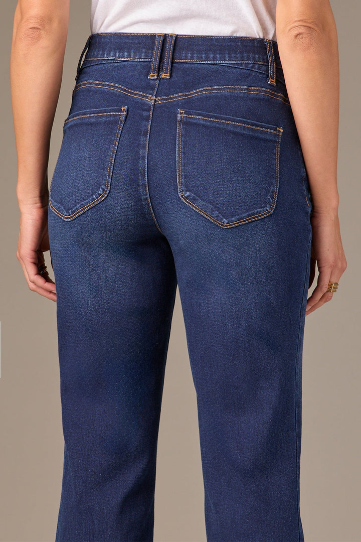 Rear view of a person wearing dark blue denim jeans with a white top, showing the back pockets and waistband details against a solid beige background.