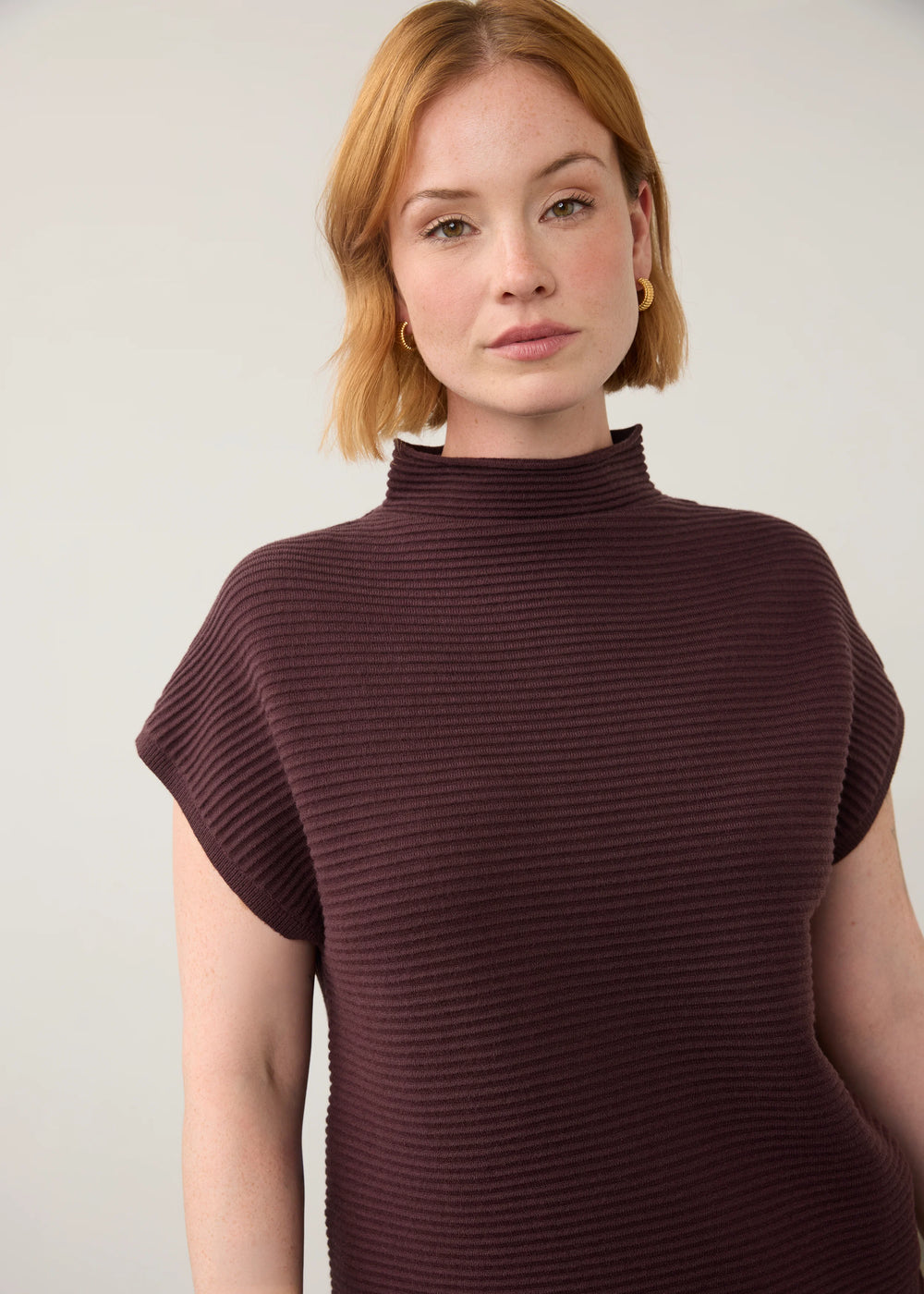 A woman with short blonde hair wearing a ribbed, short-sleeve mock neck top in a deep plum-brown color, photographed against a plain light gray background.