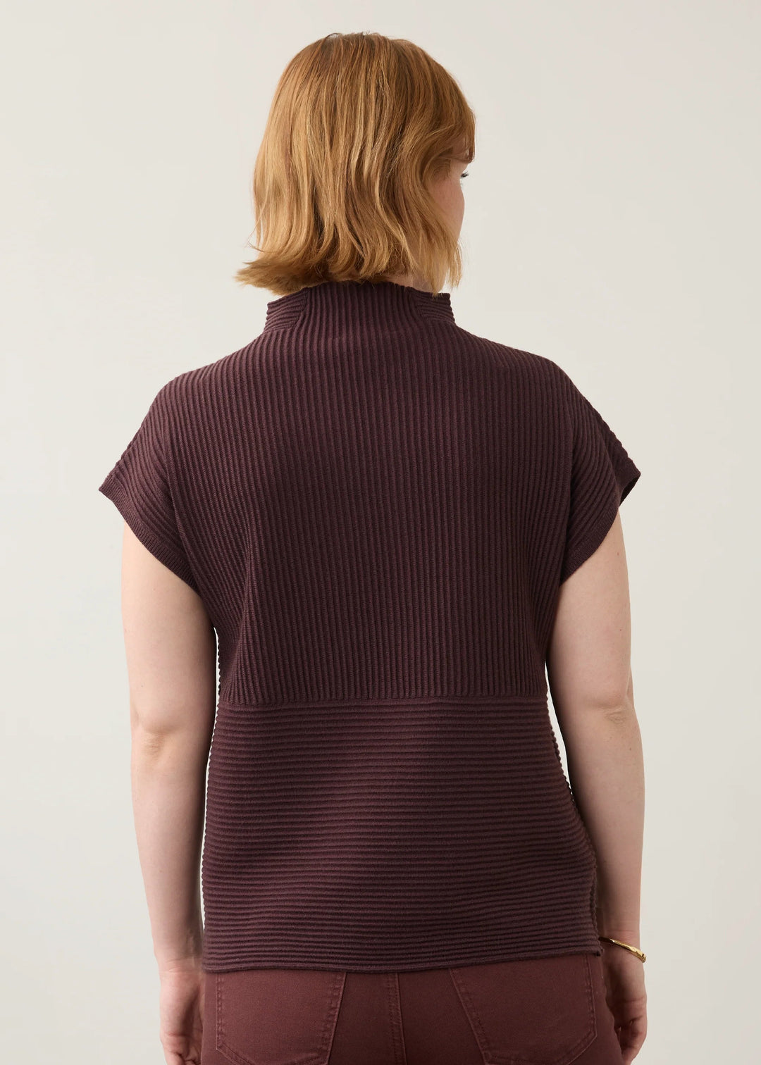 A woman viewed from the back wearing a short-sleeve ribbed mock-neck top and matching ribbed trousers in a deep burgundy color, set against a plain light gray studio background.