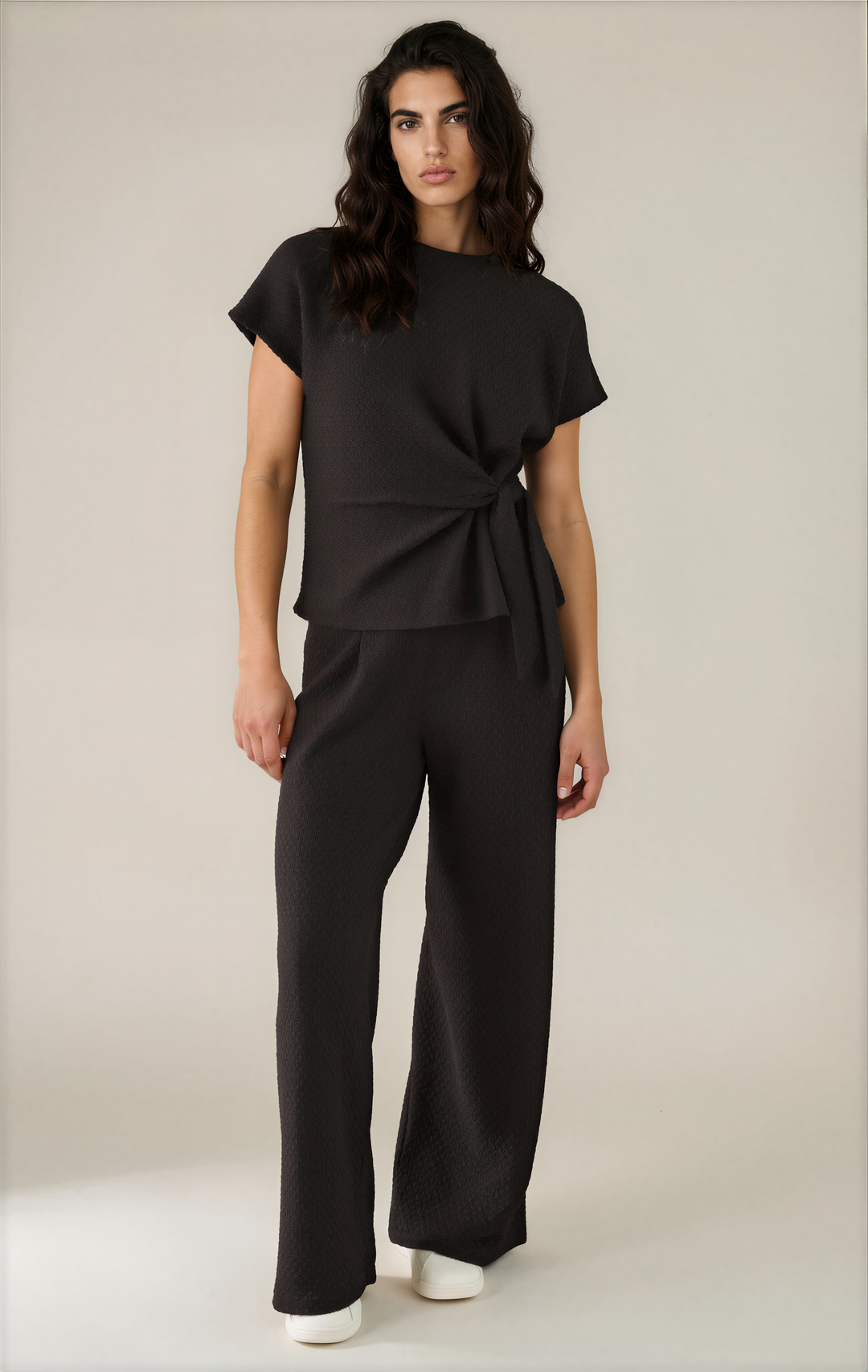 A woman modeling a black two-piece outfit consisting of a short-sleeve wrap-style top and matching wide-leg trousers, photographed against a plain light beige studio background.