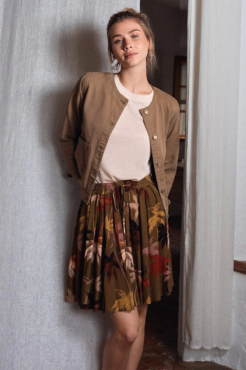 Woman wearing a brown jacket, white shirt, and floral skirt standing against a textured wall.