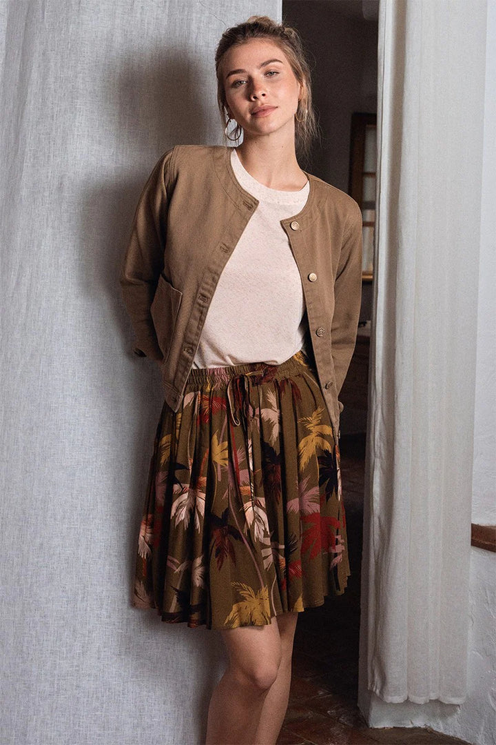 Woman wearing a brown jacket, white shirt, and floral skirt standing against a textured wall.