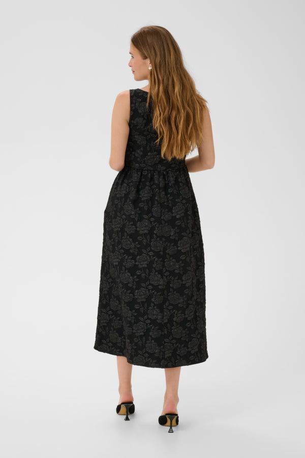 A woman with long light brown hair is shown from the back wearing a sleeveless black midi dress with a subtle tonal floral jacquard pattern. She is wearing black and gold pointed-toe mule heels against a plain light gray studio background.