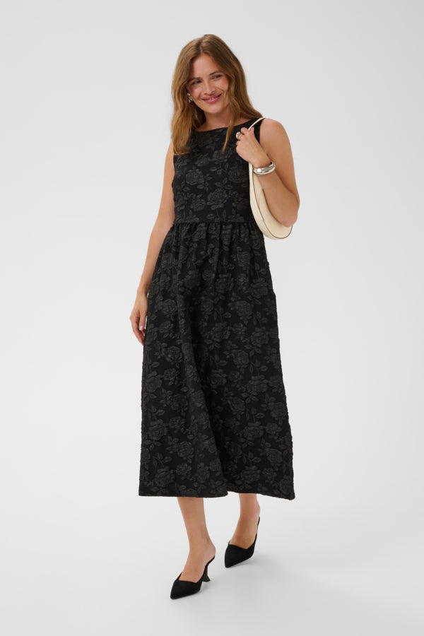 A woman modeling a sleeveless black midi dress with a subtle tonal floral jacquard pattern, paired with black pointed-toe heels and a cream shoulder bag, set against a plain light gray studio background.