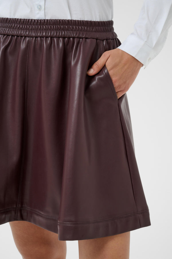 A close-up of a person wearing a dark brown faux-leather skirt with an elastic waistband and side pockets, paired with a white button-up shirt against a light gray background.