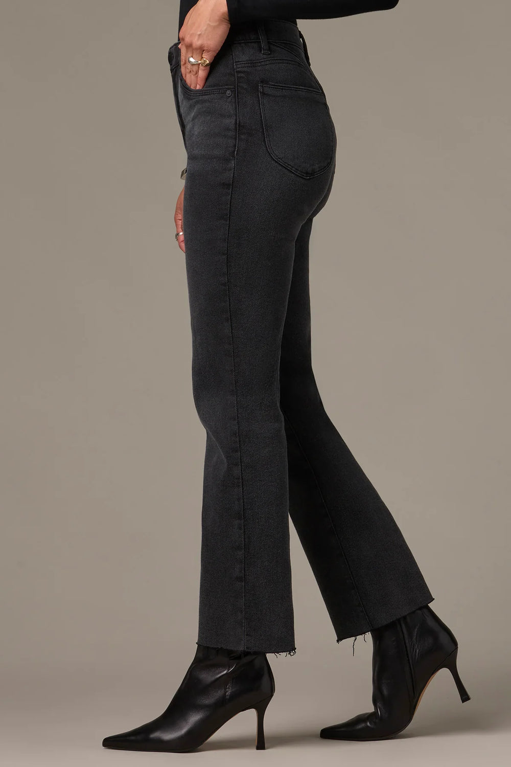 A model wearing a pair of dark charcoal gray flare jeans with raw frayed hems, paired with black pointed-toe heeled ankle boots, shown from a side profile against a solid taupe background.
