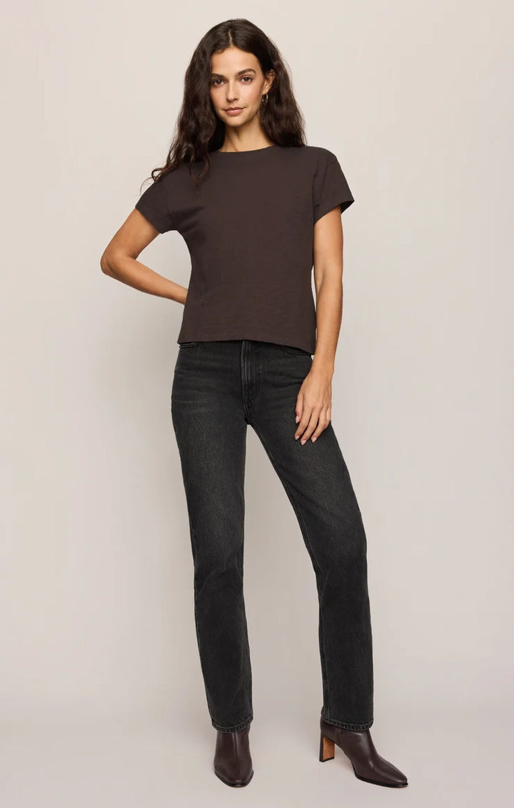 A woman modeling a dark brown short-sleeve crewneck t-shirt and dark wash straight-leg jeans, paired with dark brown heeled ankle boots, against a light gray studio background.