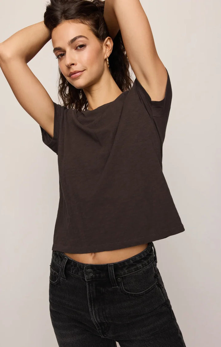 A woman modeling a dark brown cropped t-shirt with a wide draped neckline, paired with black jeans, against a plain light gray background.