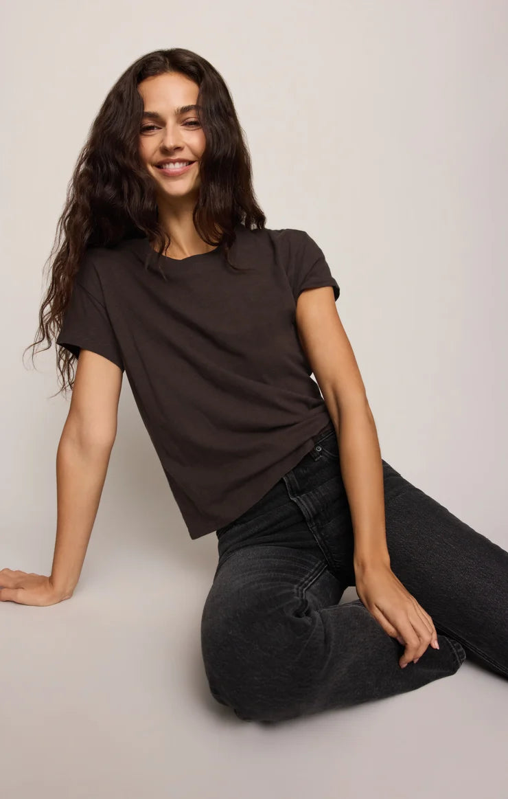 A woman with long dark wavy hair sits on a light gray floor against a light gray wall, wearing a dark brown short-sleeve crewneck t-shirt and dark gray jeans.