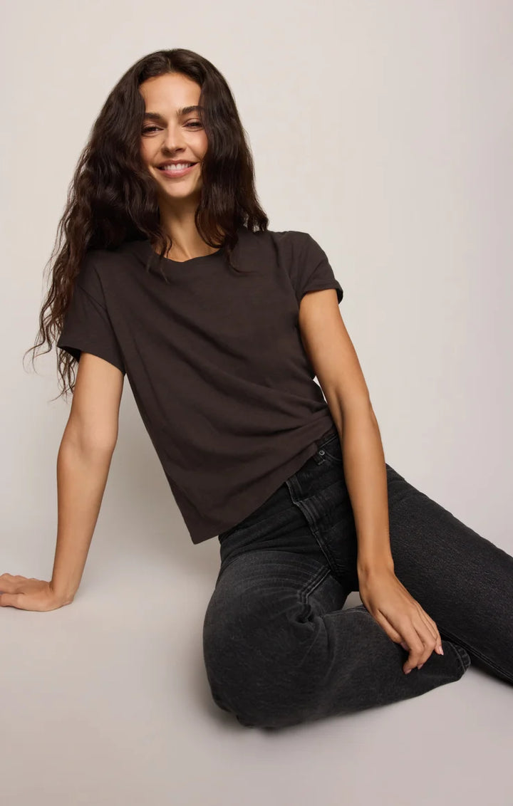 A woman with long dark wavy hair sits on a light gray floor against a light gray wall, wearing a dark brown short-sleeve crewneck t-shirt and dark gray jeans.