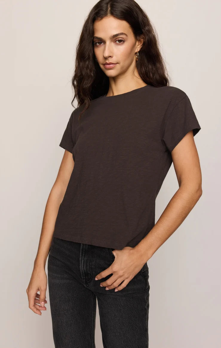 A woman modeling a dark brown short-sleeve t-shirt with a wide boat neckline, paired with black jeans, against a plain light gray background.