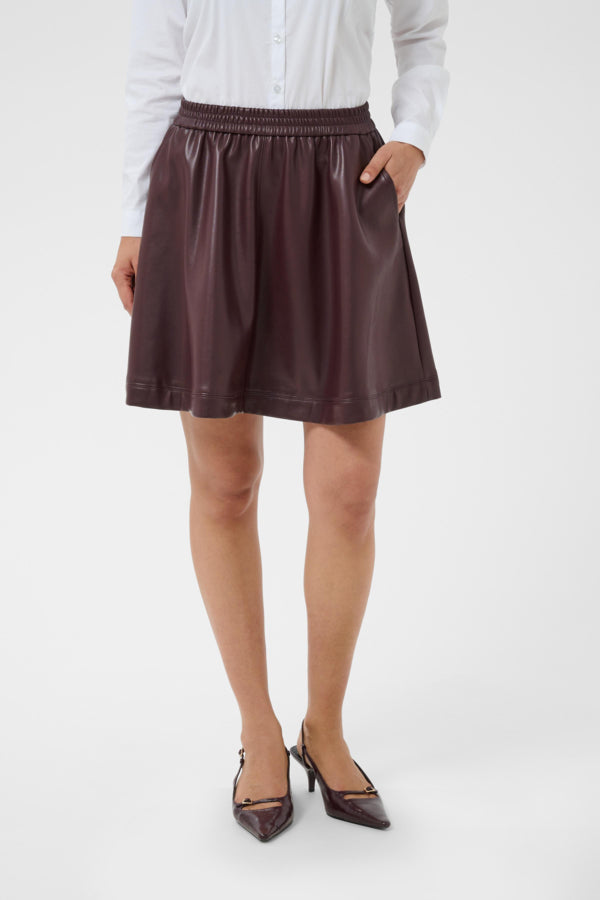 A model wearing a dark brown faux-leather mini skirt with an elastic waistband, paired with a white long-sleeve blouse and dark brown pointed-toe heels, against a plain white studio background.