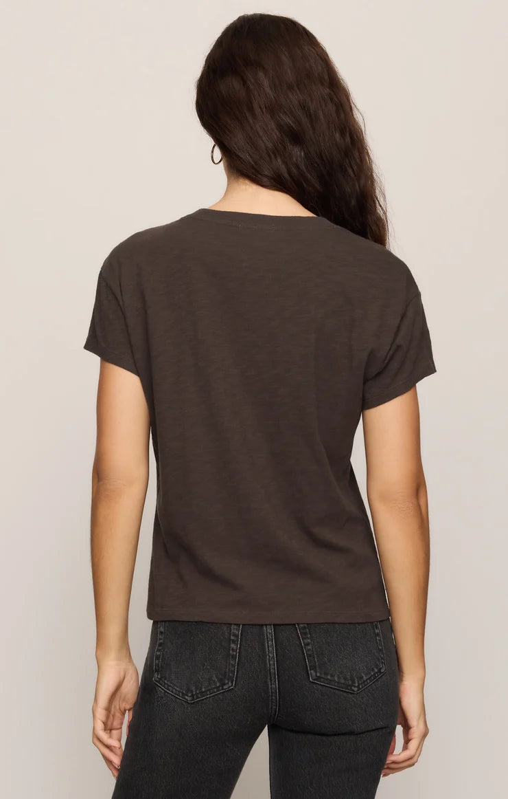 A woman viewed from the back wearing a dark brown short-sleeve crewneck t-shirt and dark gray jeans, set against a plain light gray studio background.