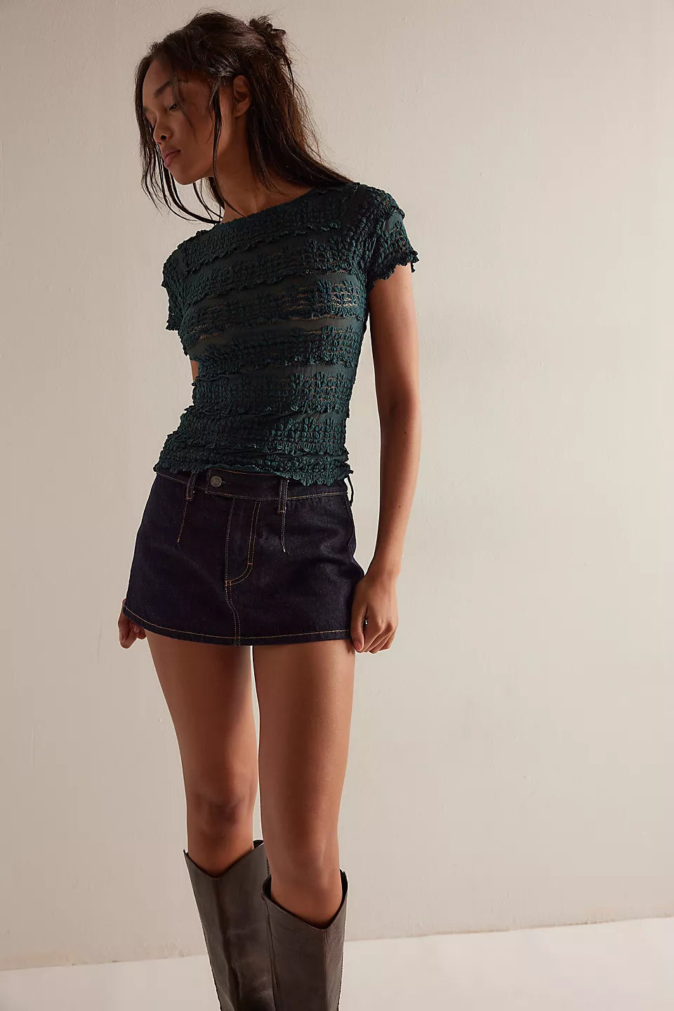 Woman wearing a dark green textured top and black shorts with gray boots on a plain background
