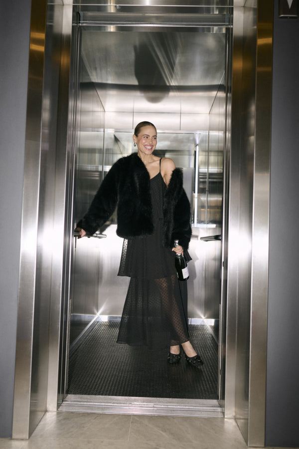 A woman stands inside a metallic elevator lobby wearing a black fur jacket over a black off-the-shoulder dress and holding a small black handbag.