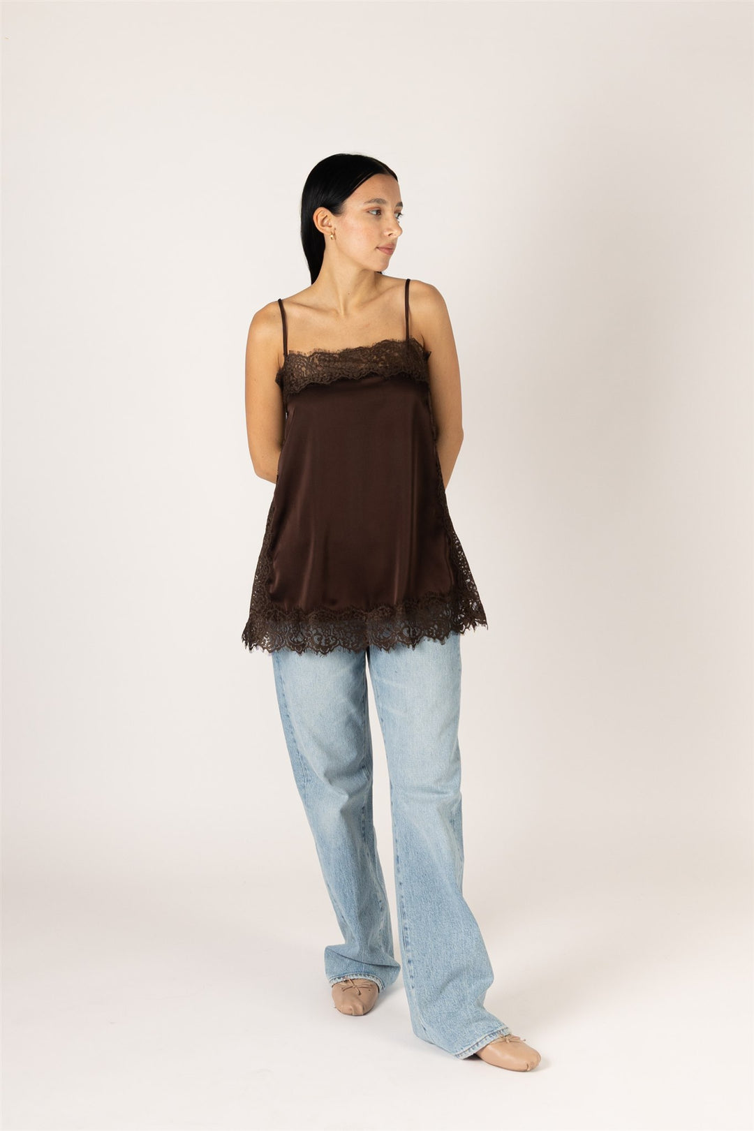 A woman modeling a dark brown satin camisole top with black lace trim, paired with light blue jeans, against a plain off-white studio background.