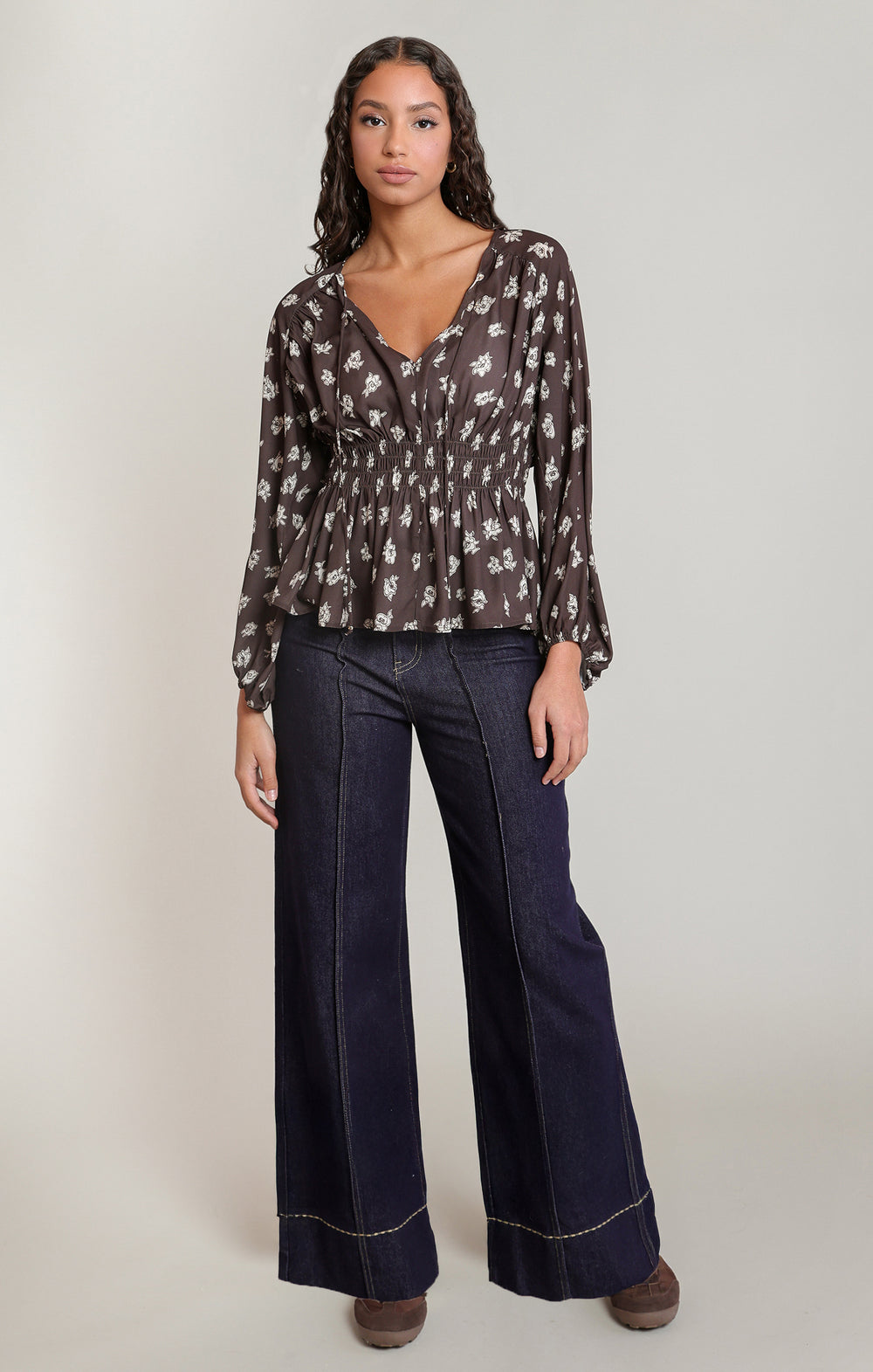 A woman modeling a sheer long-sleeve blouse with a white floral print on a dark brown background, paired with dark wide-leg jeans and brown shoes, against a light gray studio background.