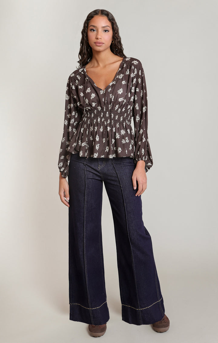 A woman modeling a sheer long-sleeve blouse with a white floral print on a dark brown background, paired with dark wide-leg jeans and brown shoes, against a light gray studio background.