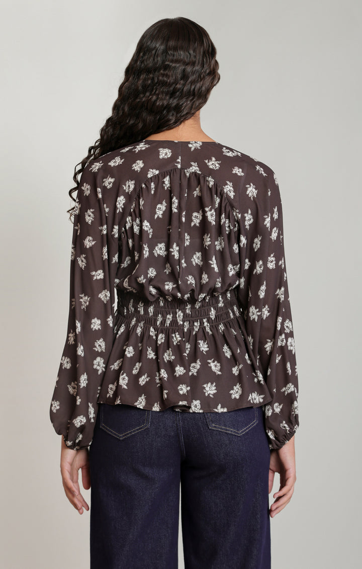 A woman shown from the back wearing a dark brown long-sleeve blouse with a white floral pattern and dark blue jeans, against a light gray studio background.
