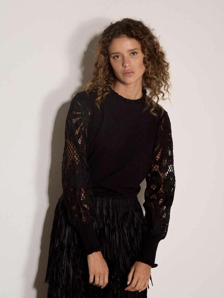 A woman with curly brown hair wearing a black long-sleeve top with sheer crochet lace sleeves and a black pleated skirt, standing against a plain off-white wall.