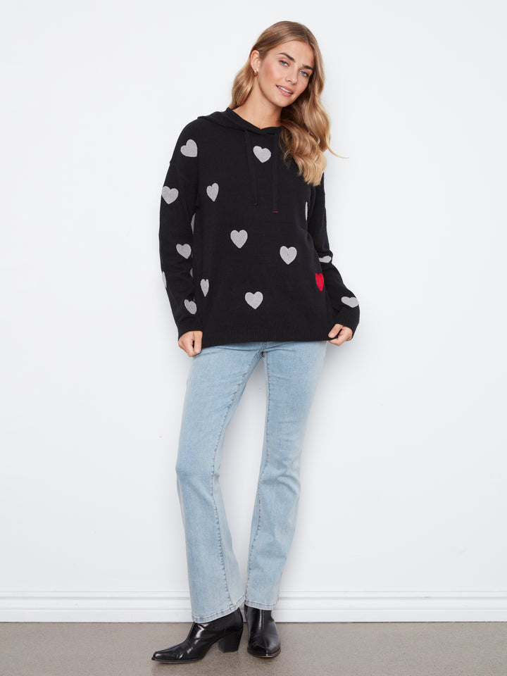 A woman modeling a black pullover hoodie with a pattern of light gray hearts and a small red accent on the pocket, paired with light-wash blue jeans and black ankle boots, standing against a white wall.