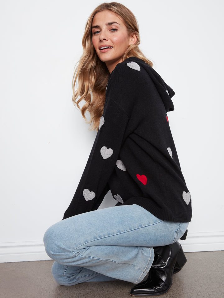 Woman wearing a black hoodie with heart patterns, sitting on the floor against a white wall.