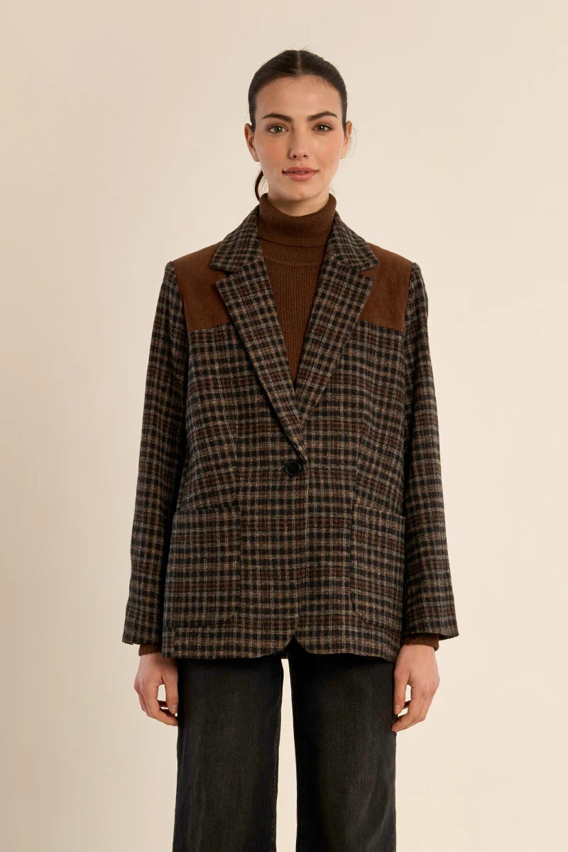 A model wearing a brown and black plaid blazer with brown suede shoulder panels, styled over a brown turtleneck and dark wide-leg jeans, against a solid beige background.