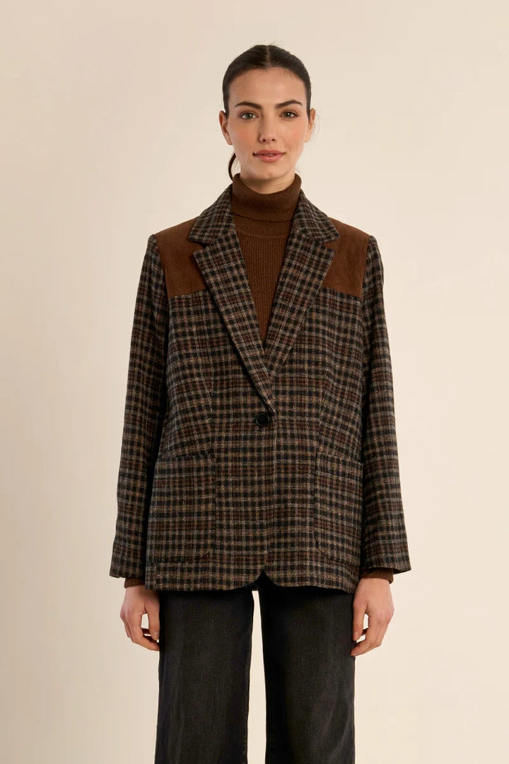 A model wearing a brown and black plaid blazer with brown suede shoulder panels, styled over a brown turtleneck and dark wide-leg jeans, against a solid beige background.