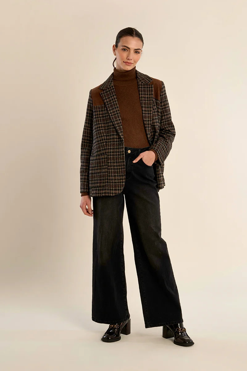 A model stands against a plain beige studio background wearing a brown and black plaid blazer with brown shoulder patches over a brown turtleneck, paired with wide-leg black jeans and black lace-up shoes.