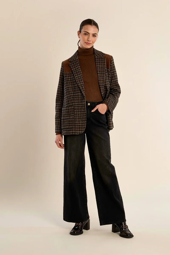 A model stands against a plain beige studio background wearing a brown and black plaid blazer with brown shoulder patches over a brown turtleneck, paired with wide-leg black jeans and black lace-up shoes.