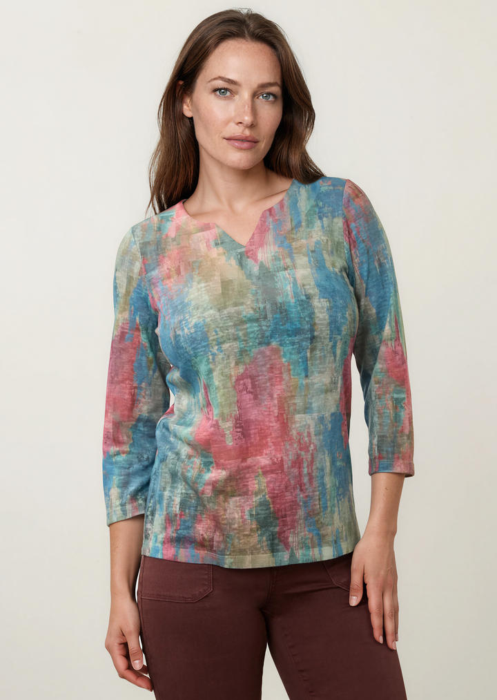A woman modeling a three-quarter sleeve V-neck top with an abstract multicolor print in shades of blue, pink, and beige, paired with dark brown pants, against a plain light gray studio background.