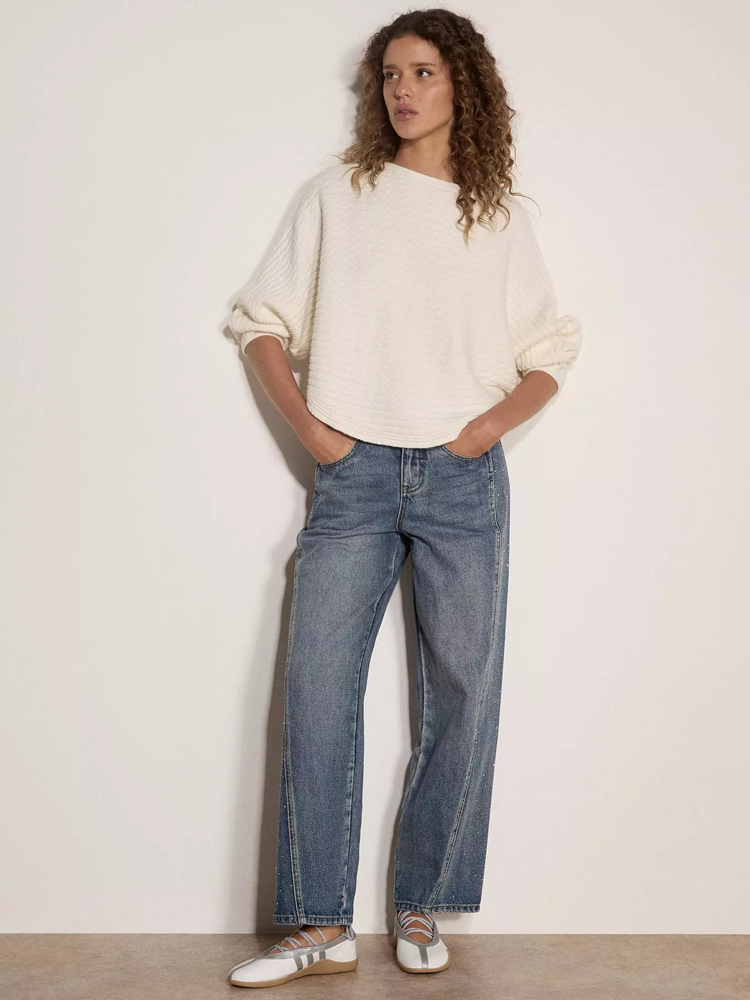 A woman stands against a plain off-white wall wearing a cream-colored textured knit sweater, blue wide-leg jeans, and white sneakers with metallic details.