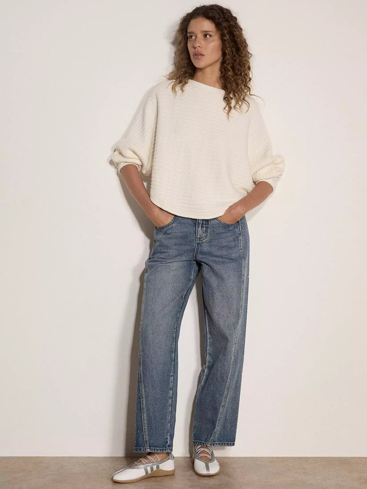 A woman stands against a plain off-white wall wearing a cream-colored textured knit sweater, blue wide-leg jeans, and white sneakers with metallic details.