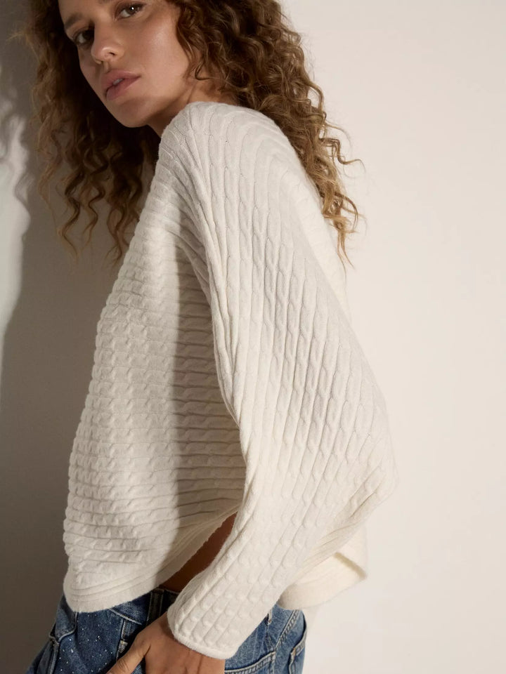 A woman wearing a cream-colored, textured knit sweater with a diamond cable pattern and draped batwing sleeves, paired with blue jeans against a neutral off-white background.