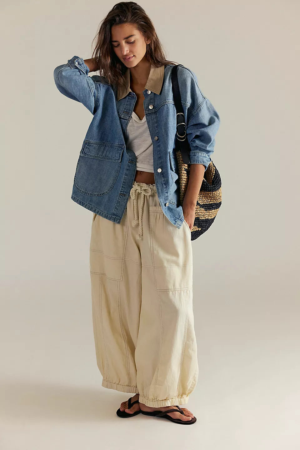 Woman wearing a denim jacket, white top, beige pants, and black sandals on a plain background