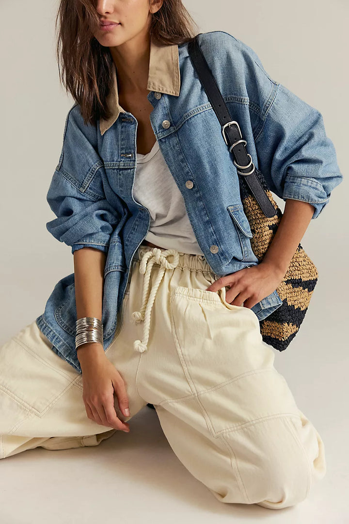 Person wearing a denim jacket and white pants with a striped bag on a neutral background