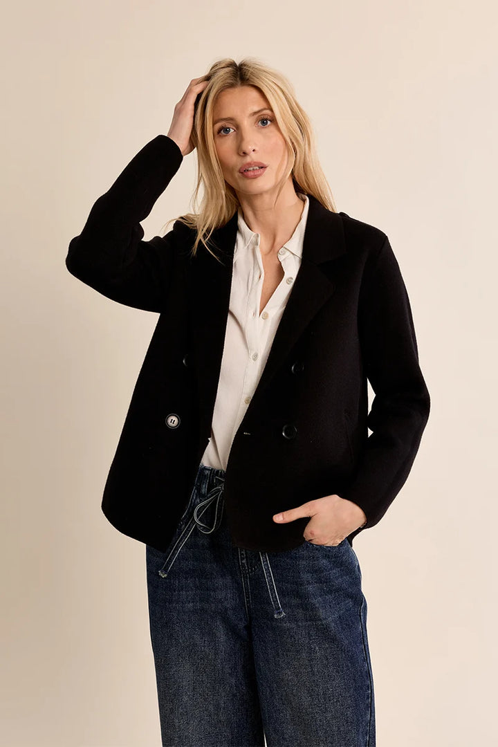 A woman modeling a black double-breasted blazer over a white button-down shirt and blue jeans, standing against a solid beige studio background.