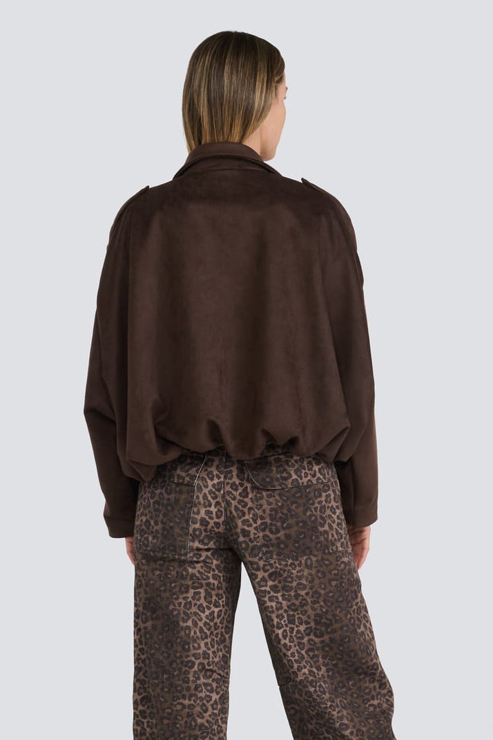 A model viewed from behind wearing a dark brown long-sleeve cropped jacket and brown leopard-print trousers, standing against a light gray studio background.