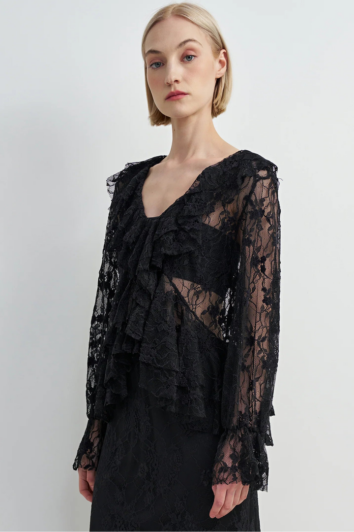 A model with short blonde hair wearing a black sheer lace ruffled blouse with long bell sleeves, paired with a black skirt, against a plain light gray studio background.