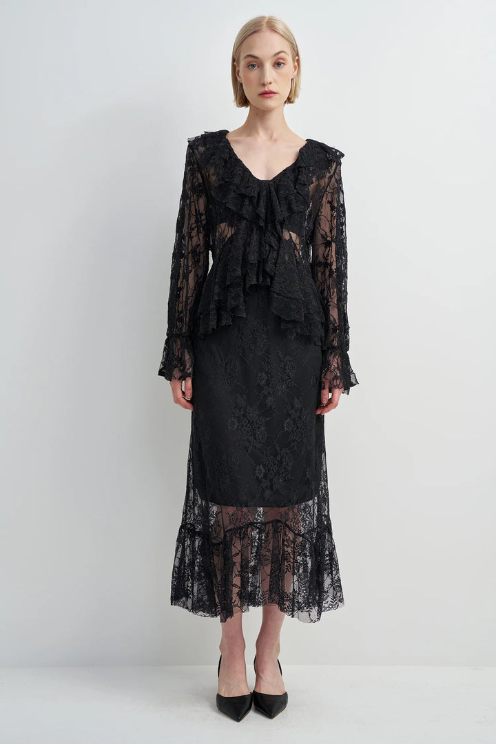 A model wearing a black lace long-sleeve top and matching midi skirt with sheer ruffle layers, paired with black pointed-toe heels, against a plain light gray studio background.