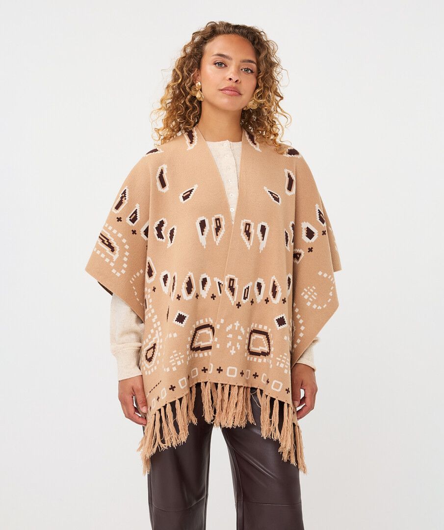 A woman modeling a beige open-front poncho with brown and white geometric patterns and fringe along the hem, worn over a white top and dark pants against a plain light gray studio background.